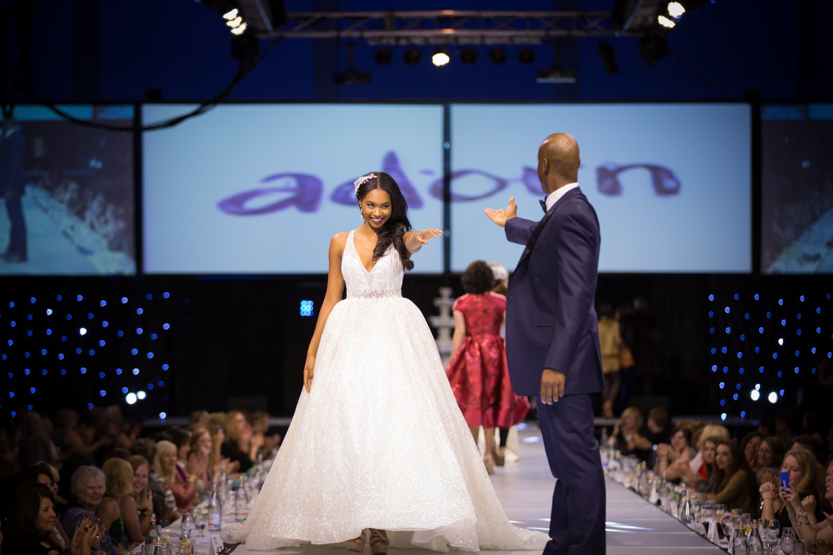 A model in a white ballgown beside a man in a navy suit on the Adorn runway.