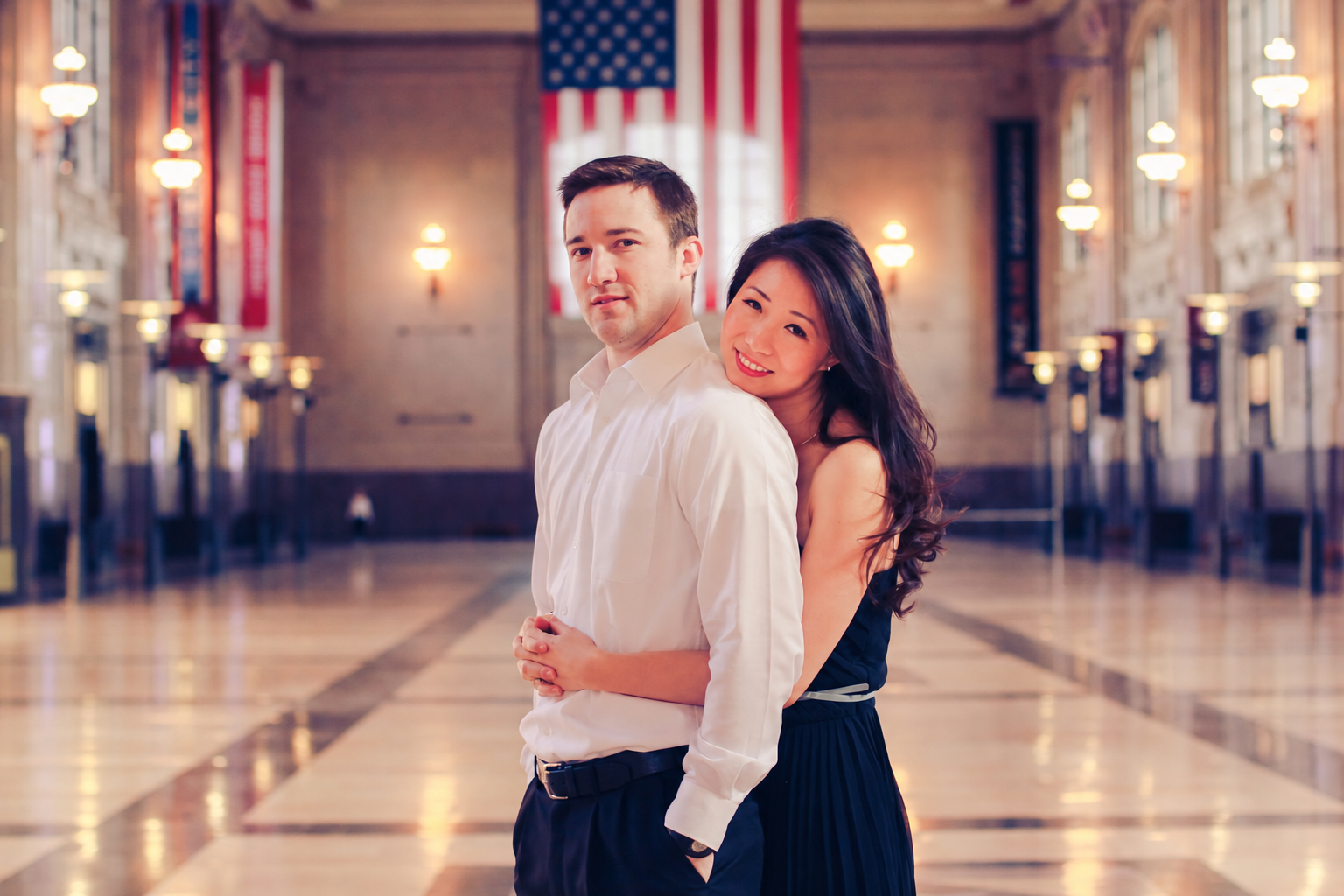 An engaged couple embracing in a bright, ornate hallway.