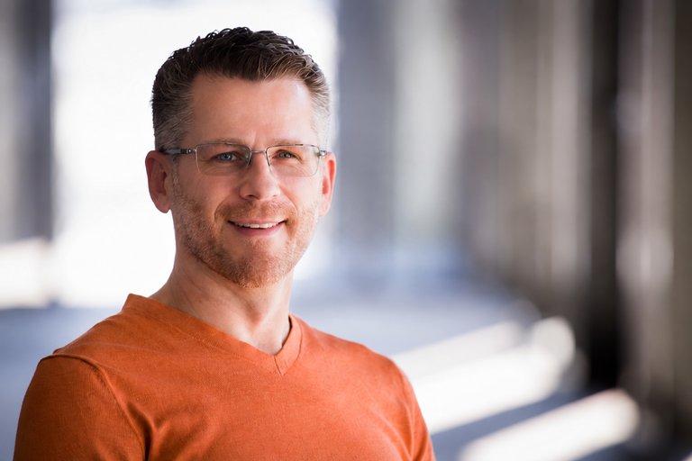 Personal brand portrait of Scott wearing glasses and an orange sweater beside softly lit windows.